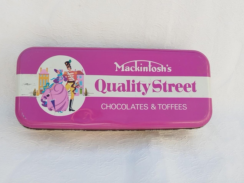 Op de afbeelding: Een roze blik met een wit label waarop "Mackintosh's Quality Street Chocolates & Toffees" staat. Het label bevat ook een cartoonafbeelding van een stel in 18e-eeuwse kleding.