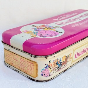 Op de afbeelding: Een vintage roze en witte blikken doos met een kleurrijke illustratie van een stel in 18e-eeuwse kleding. De tekst op de doos luidt "Mackintosh's Quality Street Chocolates".