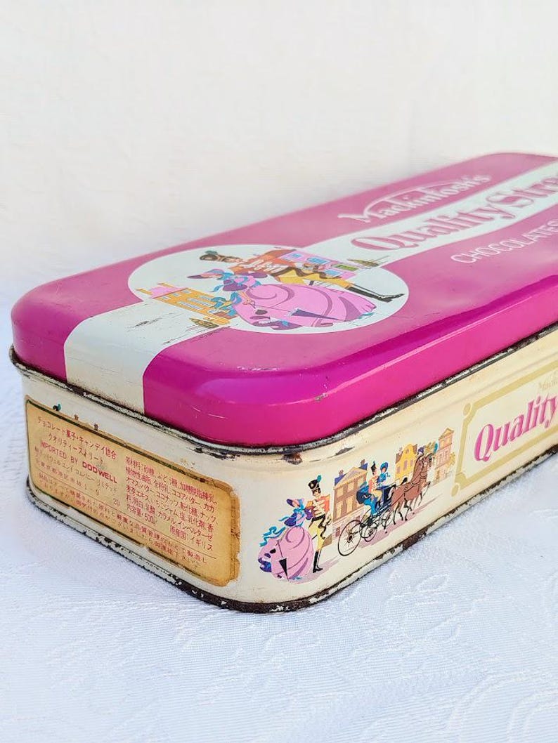 Op de afbeelding: Een vintage roze en witte blikken doos met een kleurrijke illustratie van een stel in 18e-eeuwse kleding. De tekst op de doos luidt "Mackintosh's Quality Street Chocolates".