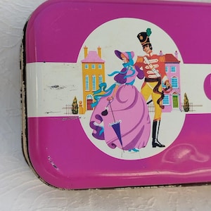 Op de afbeelding: Een roze metalen blik met een witte rand en een kleurrijke illustratie van een vrouw en een man in historische kleding. De vrouw draagt een roze jurk en een blauwe hoed. De man draagt een rood-gouden uniform. De illustratie is tegen de achtergrond van een stad met gebouwen.