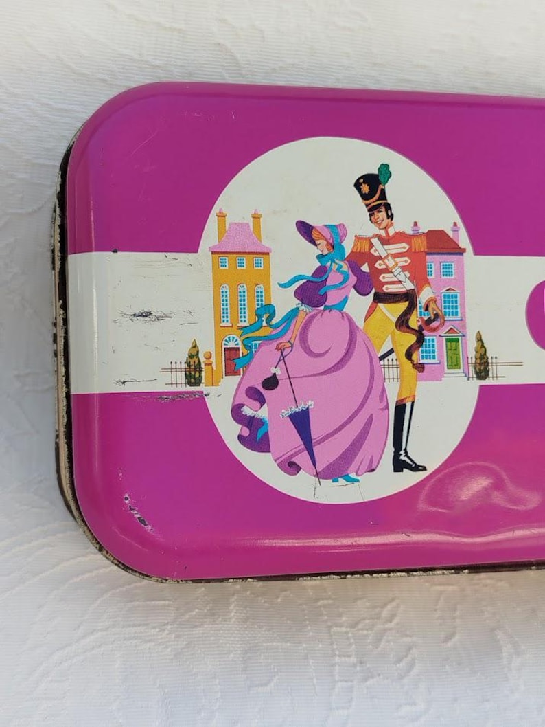 Op de afbeelding: Een roze metalen blik met een witte rand en een kleurrijke illustratie van een vrouw en een man in historische kleding. De vrouw draagt een roze jurk en een blauwe hoed. De man draagt een rood-gouden uniform. De illustratie is tegen de achtergrond van een stad met gebouwen.