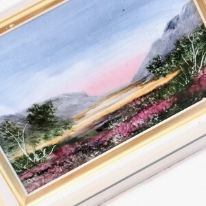May include: A framed watercolor painting depicting a landscape with a mountain range in the background, a field of purple wildflowers in the foreground, and a pink and blue sky.