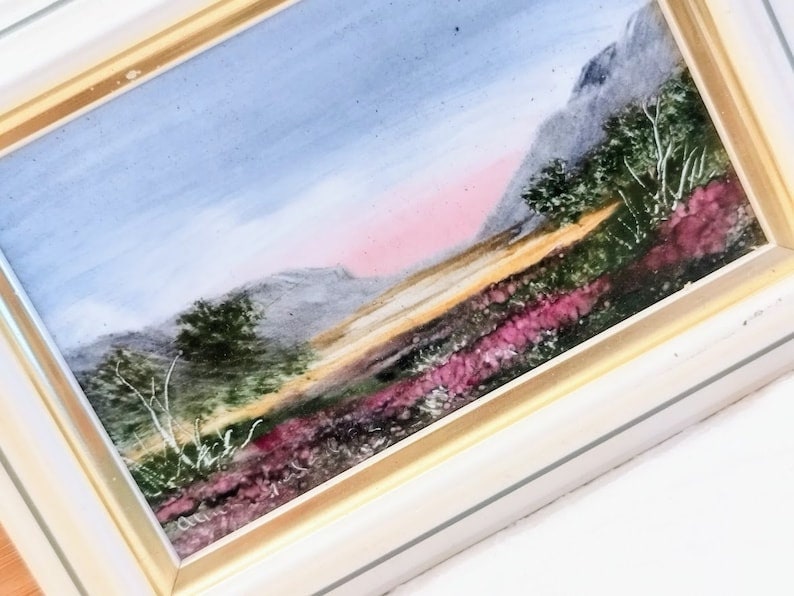 May include: A framed watercolor painting depicting a landscape with a mountain range in the background, a field of purple wildflowers in the foreground, and a pink and blue sky.