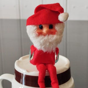 May include: A vintage Christmas decoration featuring a small Santa Claus figure. The Santa has a red felt hat, suit, and legs, with a white beard and a smiling face. The figure is perched on a white and brown mug.