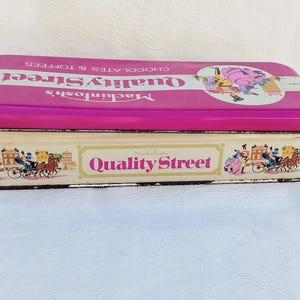Op de afbeelding: Een vintage blik voor Mackintosh's Quality Street-chocolaatjes. De doos is roze met witte letters en heeft een kleurrijke illustratie van mensen die in een koets rijden. De onderkant van de doos heeft een witte achtergrond met de woorden "Mackintosh's Quality Street" in roze letters.