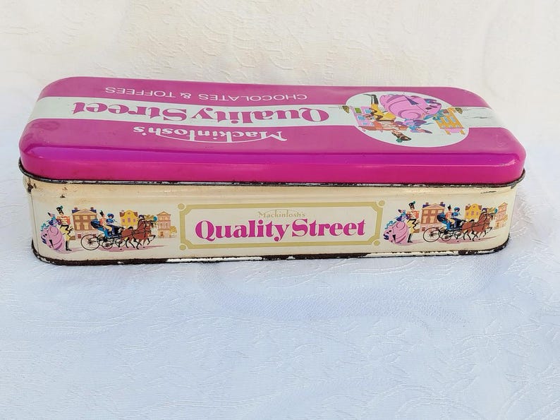 Op de afbeelding: Een vintage blik voor Mackintosh's Quality Street-chocolaatjes. De doos is roze met witte letters en heeft een kleurrijke illustratie van mensen die in een koets rijden. De onderkant van de doos heeft een witte achtergrond met de woorden "Mackintosh's Quality Street" in roze letters.