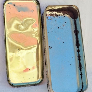 Op de afbeelding: Twee vintage rechthoekige metalen dozen met deksels. De bovenkant van de ene doos is goud met een versleten afwerking. De bovenkant van de andere doos is wit met een blauwe binnenkant en een versleten afwerking.