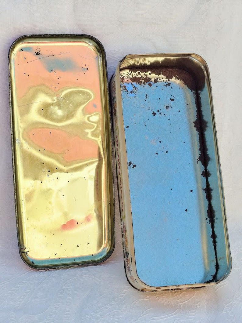 Op de afbeelding: Twee vintage rechthoekige metalen dozen met deksels. De bovenkant van de ene doos is goud met een versleten afwerking. De bovenkant van de andere doos is wit met een blauwe binnenkant en een versleten afwerking.