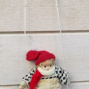 Puede incluir: Mu&ntilde;eca hecha a mano en un columpio de madera. La mu&ntilde;eca lleva un gorro, bufanda y calcetines rojos, un su&eacute;ter blanco y negro y pantalones beige. El columpio est&aacute; suspendido por cuerdas blancas.