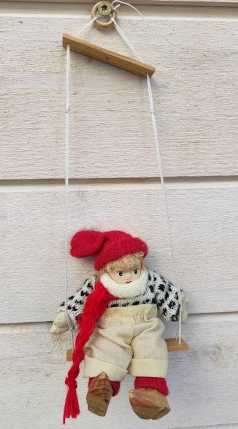 Puede incluir: Mu&ntilde;eca hecha a mano en un columpio de madera. La mu&ntilde;eca lleva un gorro, bufanda y calcetines rojos, un su&eacute;ter blanco y negro y pantalones beige. El columpio est&aacute; suspendido por cuerdas blancas.