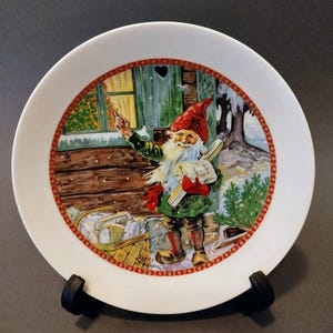 May include: A white ceramic plate featuring a festive illustration of a gnome in a red hat and green coat, holding letters. The plate has a red and gold border and depicts a snowy scene with a wooden cabin and a basket of mail.