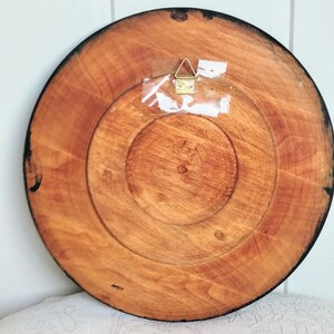 May include: Back of a round wooden decorative plate with a dark brown rim. The plate has a natural wood grain pattern and a metal hanger attached with clear tape. The plate is approximately 10 inches in diameter.