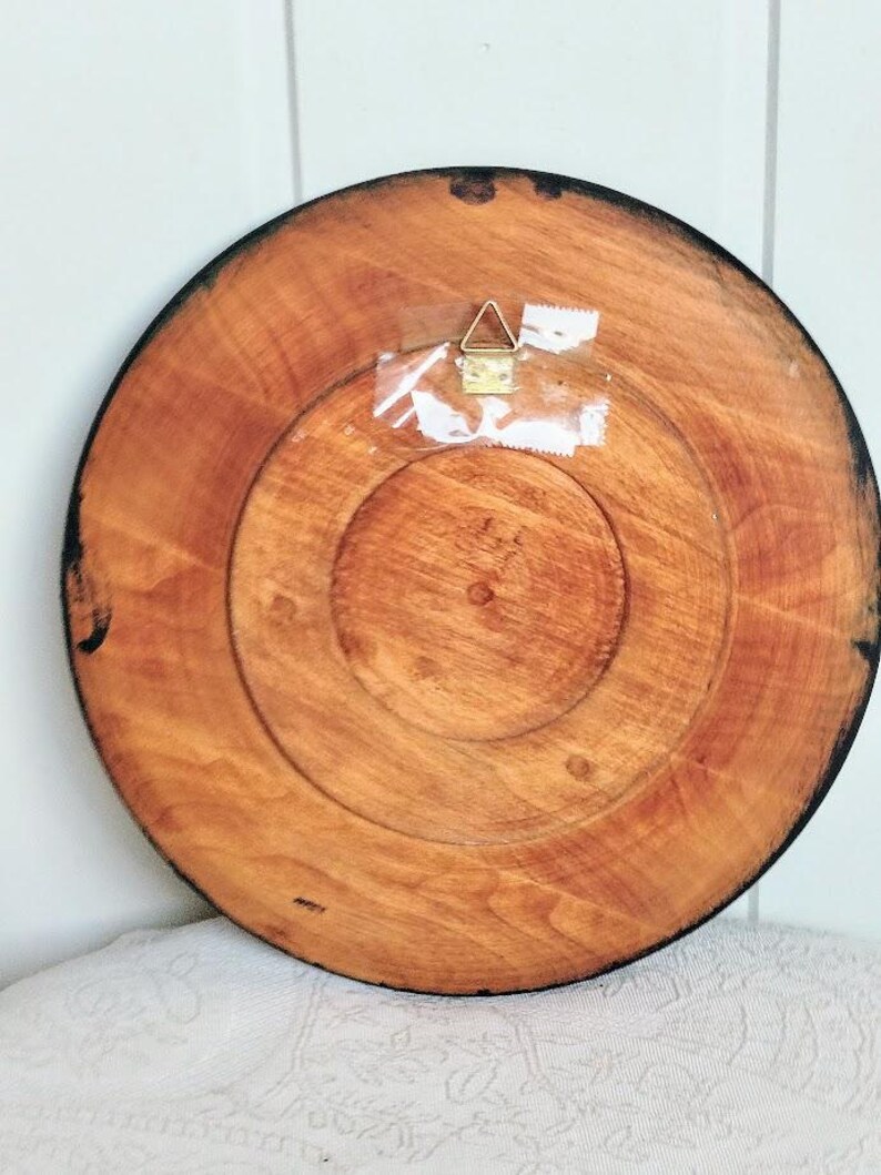 May include: Back of a round wooden decorative plate with a dark brown rim. The plate has a natural wood grain pattern and a metal hanger attached with clear tape. The plate is approximately 10 inches in diameter.