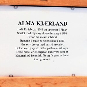 May include: A framed plaque with black text on a white background. The text reads "ALMA KJ&AElig;RLAND" and describes the artist's life and work. The plaque is made of wood and has a light brown finish.