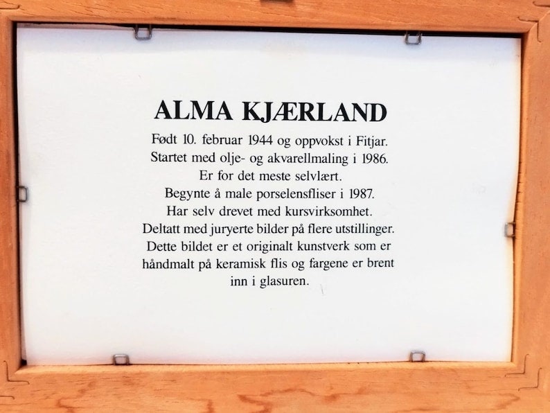 May include: A framed plaque with black text on a white background. The text reads "ALMA KJ&AElig;RLAND" and describes the artist's life and work. The plaque is made of wood and has a light brown finish.