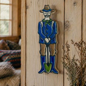 Puede incluir: Decoraci&oacute;n de pared de madera que representa a un jardinero. La figura viste una chaqueta azul, pantalones marrones y botas azules, sosteniendo una pala. El sombrero y las botas son marrones. Montado en una pared de madera.