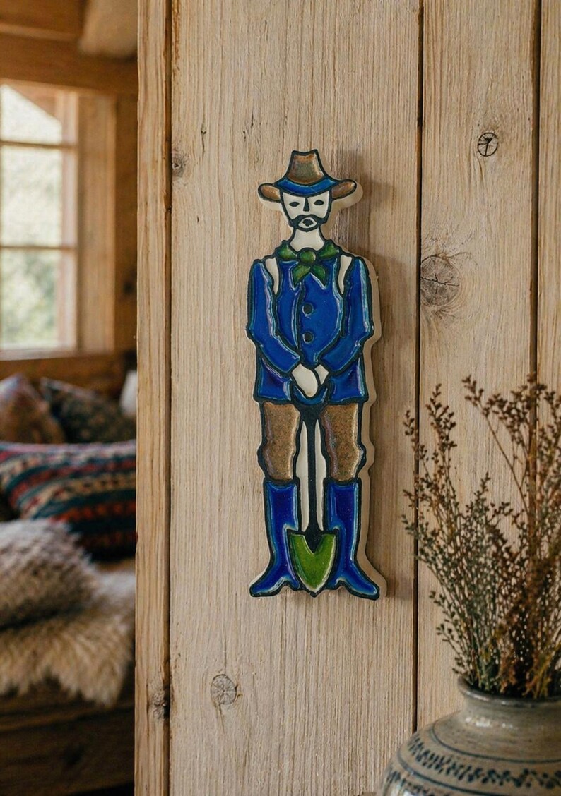 Puede incluir: Decoraci&oacute;n de pared de madera que representa a un jardinero. La figura viste una chaqueta azul, pantalones marrones y botas azules, sosteniendo una pala. El sombrero y las botas son marrones. Montado en una pared de madera.