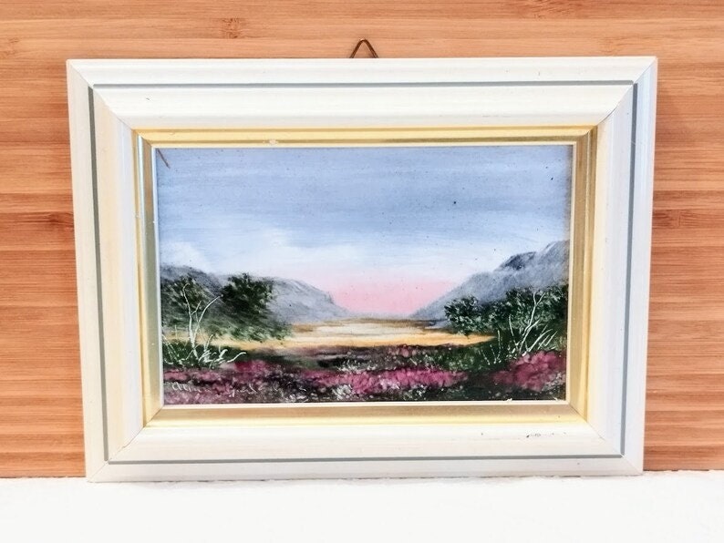 May include: A framed painting of a landscape with mountains, a lake, and purple flowers in the foreground. The sky is a light blue with a pink sunset in the distance.