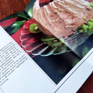 Puede incluir: Un postre rosa y blanco con relleno de frambuesa, cubierto con frambuesas frescas y rodajas de kiwi. El postre est&aacute; en un plato blanco con un borde plateado.