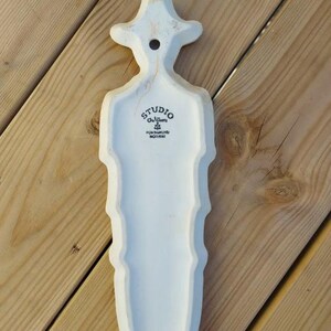 Puede incluir: Un colgante de pared de cer&aacute;mica blanca con una forma estilizada, posiblemente una pieza decorativa. Tiene una parte superior en forma de sombrero y un cuerpo largo y texturizado. El texto "STUDIO Gulbrandsen FORSGRUNN NORWAY" est&aacute; impreso.