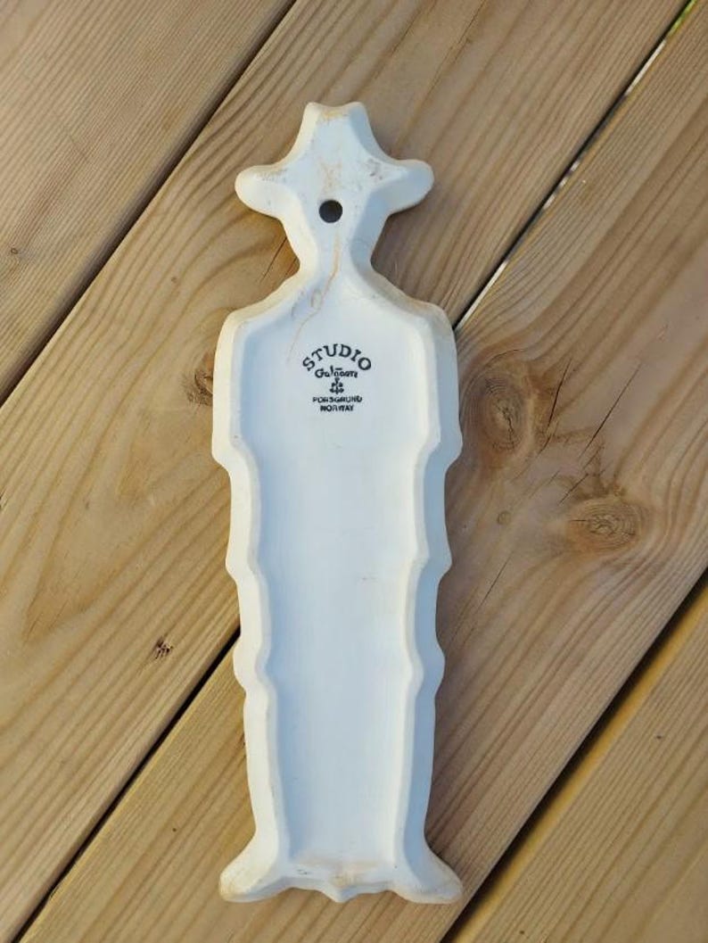 Puede incluir: Un colgante de pared de cer&aacute;mica blanca con una forma estilizada, posiblemente una pieza decorativa. Tiene una parte superior en forma de sombrero y un cuerpo largo y texturizado. El texto "STUDIO Gulbrandsen FORSGRUNN NORWAY" est&aacute; impreso.