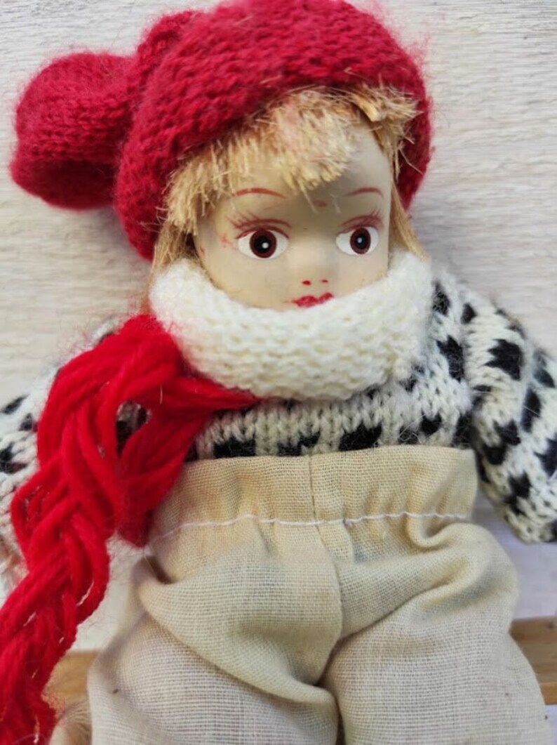 Puede incluir: Una mu&ntilde;eca vintage con la cara pintada, que lleva un gorro rojo de punto, una bufanda blanca y un jersey con estampado en blanco y negro. La mu&ntilde;eca tambi&eacute;n tiene una bufanda de hilo rojo y pantalones color beige. Un objeto de colecci&oacute;n encantador.