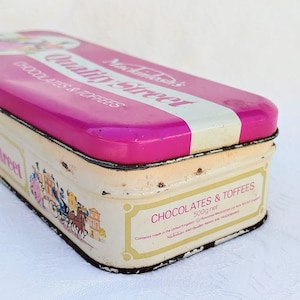 Op de afbeelding: Een vintage blik voor Quality Street chocolaatjes en toffee. De doos is roze met witte letters en een afbeelding van een koets met paarden. De tekst op de doos luidt "Mochainhasan's Quality Street Chocolates & Toffees 500g net. Contents made in the United Kingdom. "Mochainhasan's" and "Quality Street" are trademarks."