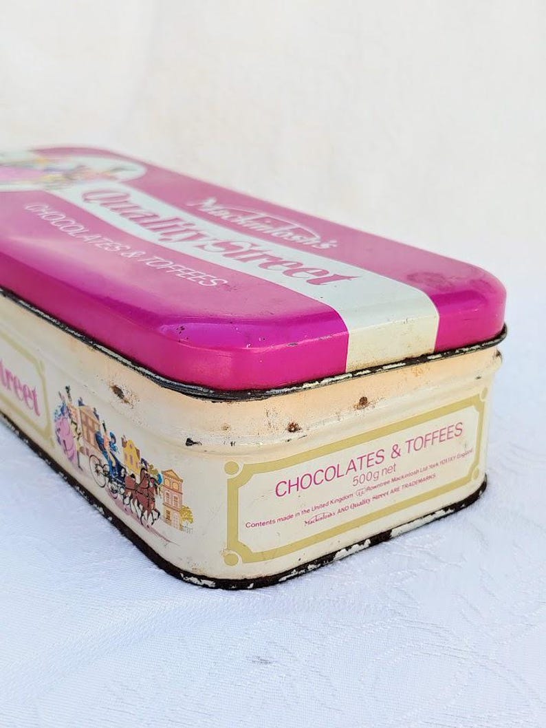 Op de afbeelding: Een vintage blik voor Quality Street chocolaatjes en toffee. De doos is roze met witte letters en een afbeelding van een koets met paarden. De tekst op de doos luidt "Mochainhasan's Quality Street Chocolates & Toffees 500g net. Contents made in the United Kingdom. "Mochainhasan's" and "Quality Street" are trademarks."