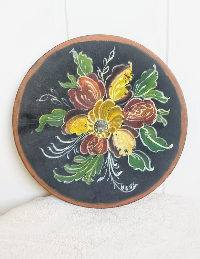 May include: Round, hand-painted wooden plate with a black background and a floral design. The flowers are in shades of yellow, brown, and red, with green leaves and white accents. The plate has a brown rim and the artist's signature.