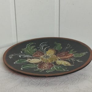 May include: A round, dark brown decorative plate with a floral design. The flowers are painted in shades of yellow, red, and green, with white accents. The plate has a dark brown rim and sits on a white patterned surface.
