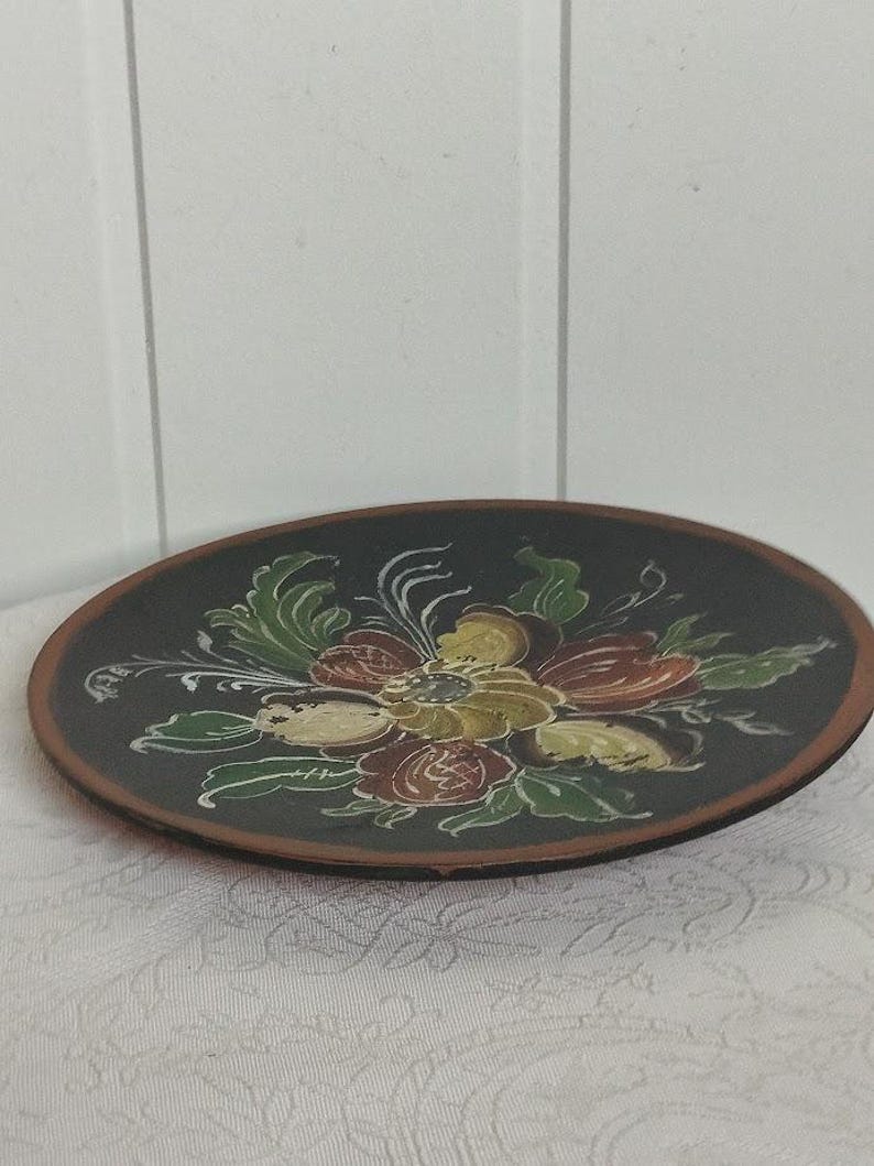 May include: A round, dark brown decorative plate with a floral design. The flowers are painted in shades of yellow, red, and green, with white accents. The plate has a dark brown rim and sits on a white patterned surface.