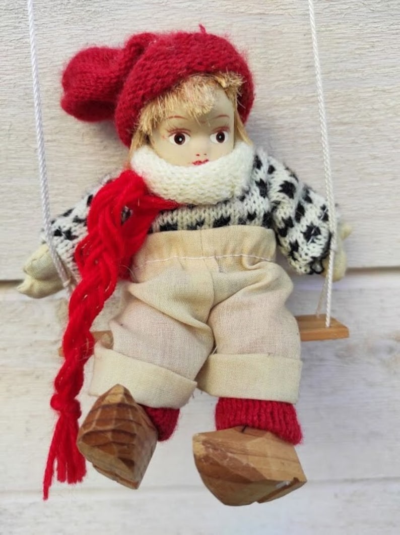 Puede incluir: Mu&ntilde;eca vintage con rostro pintado, con gorro, bufanda y calcetines de punto rojos. La mu&ntilde;eca tiene un su&eacute;ter con estampado en blanco y negro, pantalones color beige y zapatos de madera. Est&aacute; sentada en un peque&ntilde;o columpio de madera.