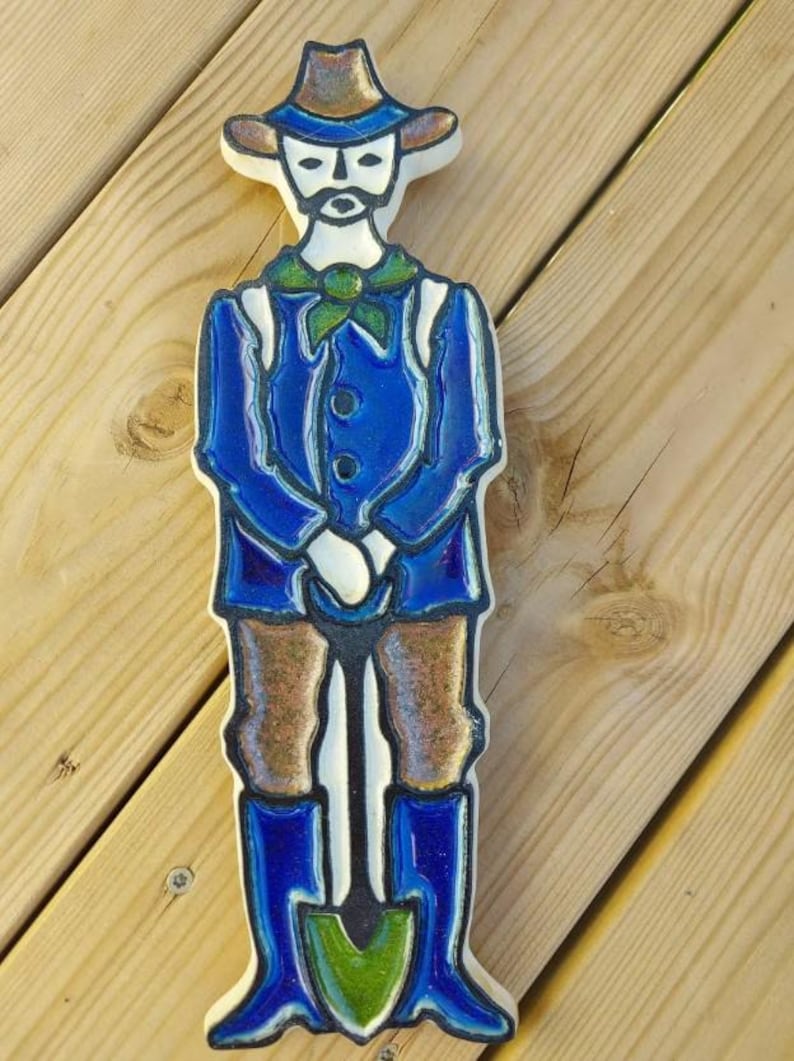 Puede incluir: Figura de cer&aacute;mica de un hombre con traje azul, pantal&oacute;n marr&oacute;n y sombrero, sosteniendo una pala. La figura tiene una cara blanca con bigote y barba. La pala tiene una hoja verde. Sobre una superficie de madera.