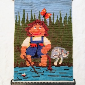May include: A vintage needlepoint wall hanging featuring a cartoon-like girl with brown hair, wearing a blue and red outfit, standing in a blue body of water with a stick in her hand. A white cat with black spots is standing next to her. A red butterfly is flying above the girl's head. The background is a green forest with a blue sky.