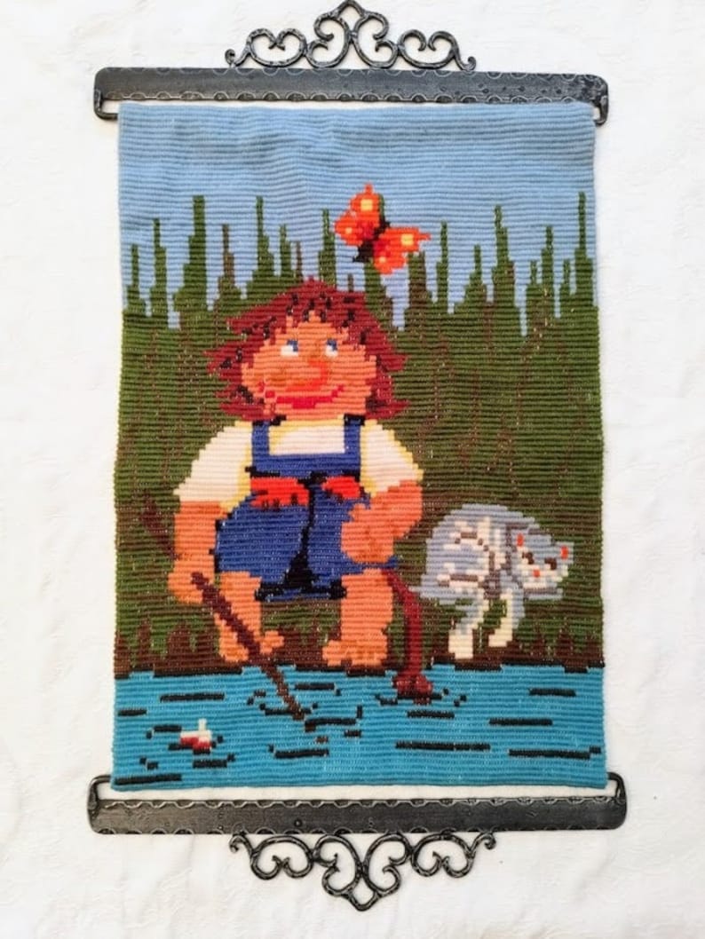 May include: A vintage needlepoint wall hanging featuring a cartoon-like girl with brown hair, wearing a blue and red outfit, standing in a blue body of water with a stick in her hand. A white cat with black spots is standing next to her. A red butterfly is flying above the girl's head. The background is a green forest with a blue sky.