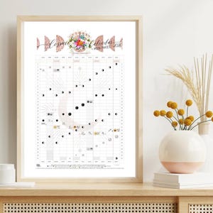 Puede incluir: Un calendario lunar enmarcado "Cosmic Calendar Moon Phases 2026". El calendario presenta una cuadrícula con ilustraciones de fases lunares y símbolos astrológicos. El título está en una fuente decorativa con detalles florales. El marco es de madera clara.