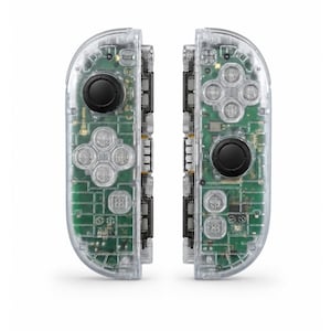 Puede incluir: Un par de mandos de videojuegos con una carcasa de plástico transparente que revela la placa de circuito verde interna, joysticks negros y botones transparentes. El mando izquierdo tiene una cruceta y el derecho cuatro botones.