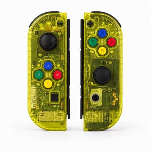 May include: A pair of neon yellow transparent game controllers featuring black joysticks and buttons. The left controller has a directional pad with blue, green, red, and yellow arrows. The right controller has four primary action buttons in blue, green, red, and yellow, plus a black plus sign button.