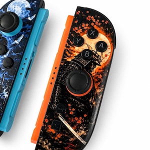 May include: Two game controllers featuring custom graphic skins. The left controller is light blue with a dark blue night sky, moon, and bird design. The right controller is orange with a black samurai, orange moon, and red maple leaf pattern.