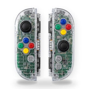 May include: A pair of clear, transparent video game controllers featuring visible internal green circuit boards. The left controller has a black joystick and four directional buttons in blue, green, red, and yellow. The right controller has a black joystick and four action buttons labeled A, B, X, and Y in red, yellow, blue, and green.