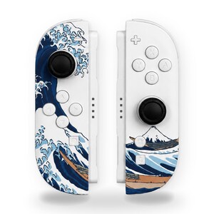 May include: Two white game controllers featuring a blue and white graphic print of a large ocean wave and a mountain. Each controller has black circular joysticks, white buttons with letters, and directional pads on a white plastic surface.