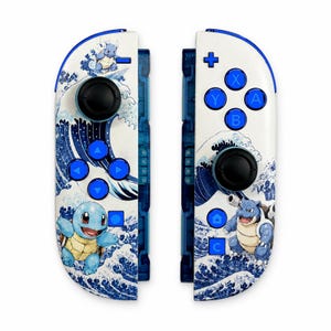 Könnte beinhalten: Ein Paar weiße Game-Controller mit blauem Wellenmuster und Illustrationen von Schiggy und Turtok. Die Controller verfügen über schwarze Joysticks, blaue Richtungstasten und blaue Aktionstasten mit A-, B-, X- und Y-Beschriftung.
