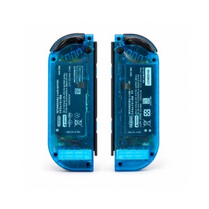 Pu&ograve; includere: Due controller di gioco blu trasparenti, ciascuno con componenti interni visibili. I controller hanno dettagli neri e includono testo che indica che sono batterie ricaricabili agli ioni di litio. Il numero del modello &egrave; HAC-006.