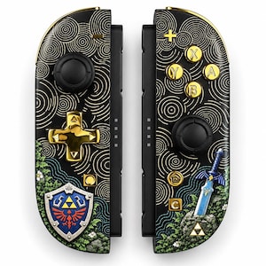 Op de afbeelding: Een paar zwarte gamecontrollers met gouden knoppen en een gouden richtingspad. Het oppervlak is versierd met gouden cirkelvormige lijnpatronen, groen gebladerte, een blauw zwaard en een schild met een rood en geel embleem op een witte achtergrond.