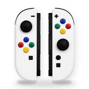 May include: A pair of white video game controllers featuring black analog sticks and a set of colorful action buttons. The left controller includes a directional pad with blue, red, green, and yellow buttons, while the right controller has blue, red, green, and yellow lettered buttons.