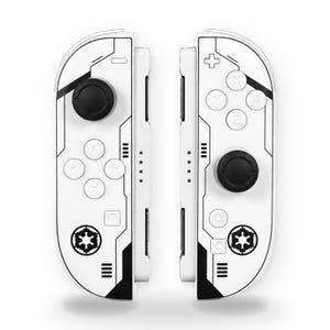 May include: A pair of white video game controllers featuring black geometric line patterns and a circular black and white Imperial crest emblem at the bottom of each unit. The controllers include black analog sticks and white buttons.