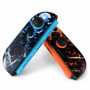 May include: Two game controllers featuring custom graphic skins. The left controller has a blue base with a moon, waves, and bird design. The right controller has an orange base with a samurai figure, sword, and red floral accents.
