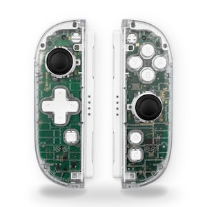 May include: A pair of video game controllers with clear plastic shells revealing the internal green circuit boards. The left controller features a black joystick and white directional pad, while the right has a black joystick and four white buttons labeled A, B, X, and Y.