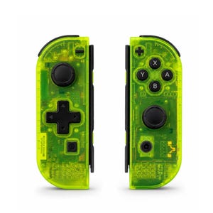 Op de afbeelding: Een paar neon-groene videogamecontrollers met een transparante behuizing die de interne elektronica zichtbaar maakt. De linkercontroller heeft een zwarte analoge stick en een D-pad, terwijl de rechtercontroller vier zwarte actieknoppen met de labels A, B, X, Y en een zwarte analoge stick bevat.