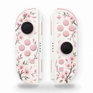 May include: A pair of white video game controllers featuring a pink cherry blossom branch pattern. Each controller has pink buttons, a black joystick, and a white base. The design includes detailed pink flowers and brown branches.